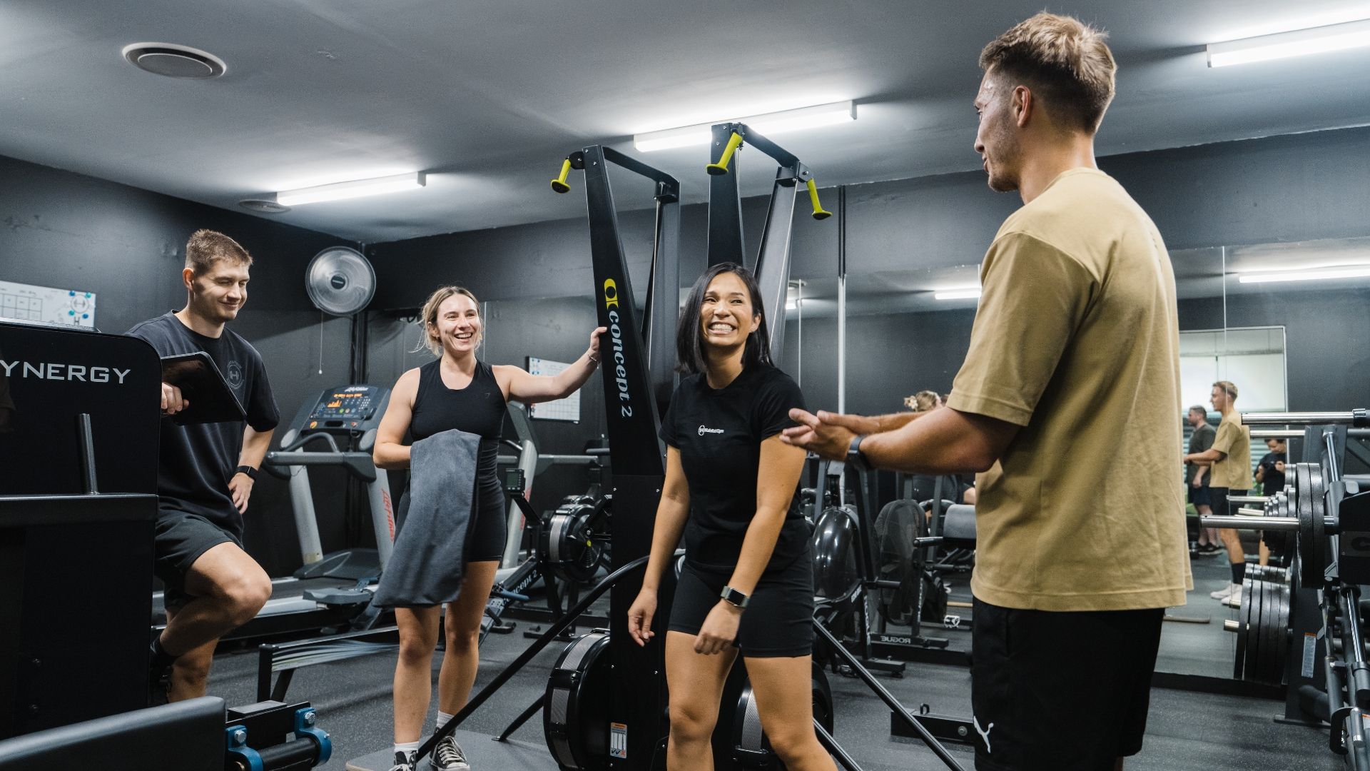 Group of personal trainers and clients smiling and chatting together in the gym, creating a positive and supportive training environment.
